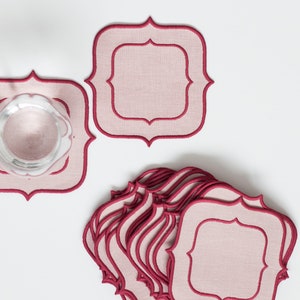 May include: A stack of pink and burgundy square fabric coasters with a decorative border. The coasters are made of linen and have a subtle texture.