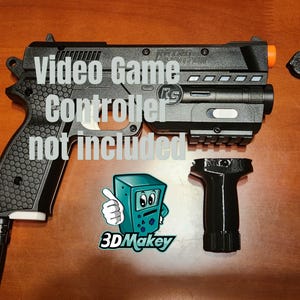 May include: A black plastic pistol-shaped accessory for a video game controller, featuring a textured honeycomb grip, an orange muzzle tip, and a detachable vertical foregrip. The text 'Video Game Controller not included' is visible.