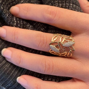 Könnte beinhalten: Ein goldener Ring mit einem filigranen Design aus Blättern und kleinen Diamanten. Der Ring ist breit und hat einen einzigartigen, durchbrochenen Stil.