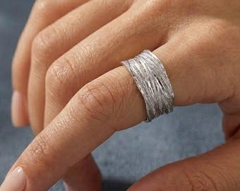 Puede incluir: Un anillo ancho de plata con un acabado cepillado y texturizado que presenta finas líneas verticales grabadas en toda la superficie. La banda tiene un ancho constante y un brillo metálico, mostrando un aspecto detallado y multicapa.