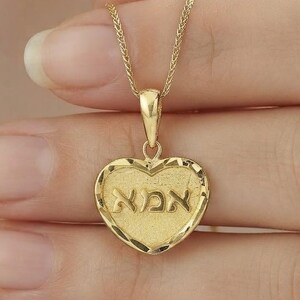 Puede incluir: Un collar con colgante en forma de coraz&oacute;n de oro con un borde texturizado y la palabra hebrea para madre, 'אמא', grabada en el centro. El colgante cuelga de una delicada cadena de oro, mostrando un acabado pulido en las letras y una superficie mate.