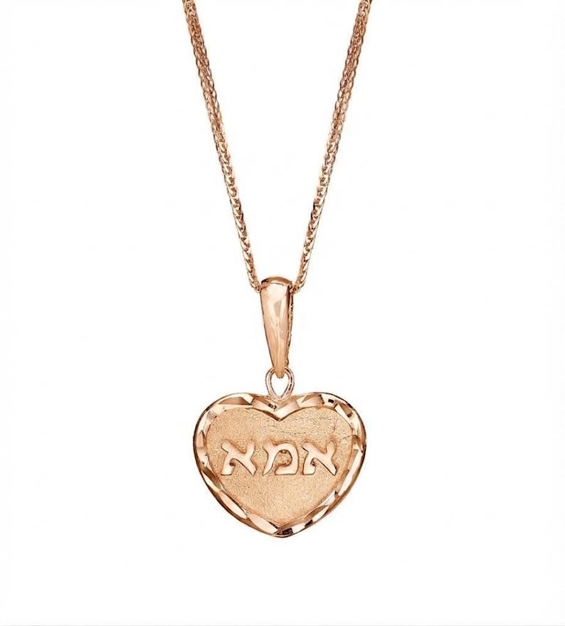 Puede incluir: Un collar con colgante en forma de coraz&oacute;n de oro rosa que presenta un centro texturizado con la palabra hebrea "אמא" grabada en el medio. El coraz&oacute;n tiene un borde facetado de corte diamante y cuelga de una delicada cadena de oro rosa.
