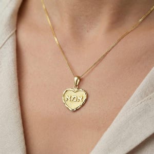 Puede incluir: Un collar con colgante en forma de coraz&oacute;n de oro, con una superficie texturizada y la palabra hebrea "אמא" grabada en el centro. El colgante cuelga de una delicada cadena de oro, mostrando un borde pulido y un dise&ntilde;o cl&aacute;sico y sencillo.