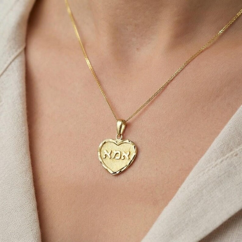 Puede incluir: Un collar con colgante en forma de coraz&oacute;n de oro, con una superficie texturizada y la palabra hebrea "אמא" grabada en el centro. El colgante cuelga de una delicada cadena de oro, mostrando un borde pulido y un dise&ntilde;o cl&aacute;sico y sencillo.