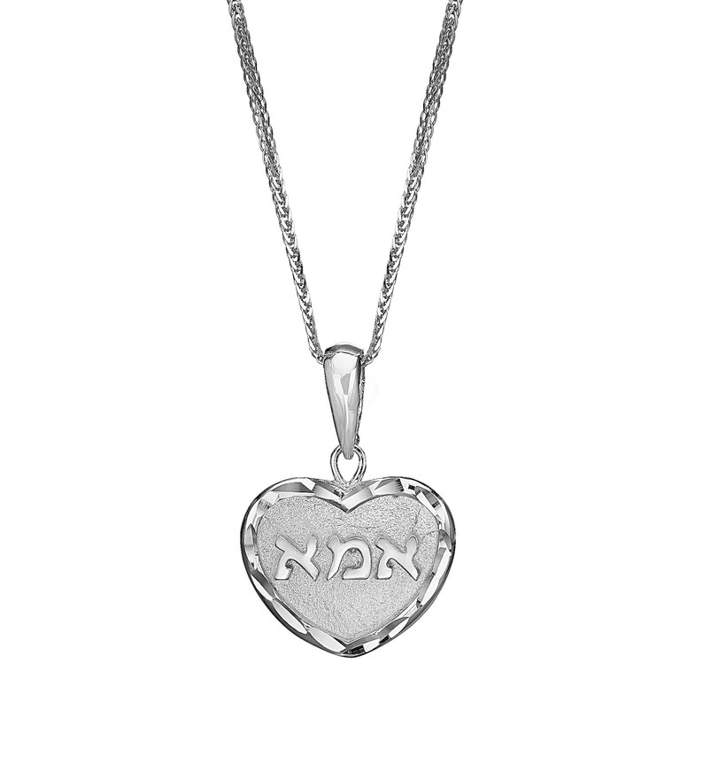 Puede incluir: Un collar con colgante en forma de coraz&oacute;n de tono plateado con un centro texturizado que tiene la palabra hebrea "אמא" grabada en el medio. El coraz&oacute;n tiene un borde pulido y facetado y cuelga de una fina cadena de eslabones plateados.