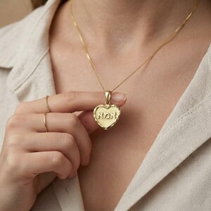 Puede incluir: Un collar con colgante en forma de coraz&oacute;n de oro que presenta la palabra hebrea אמא grabada en el frente. El colgante cuelga de una delicada cadena de oro, y se llevan dos anillos finos de oro en los dedos, a juego con el tono del metal.