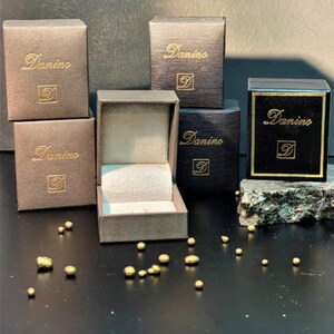 Puede incluir: Una colecci&oacute;n de cajas cuadradas para joyas en negro y marr&oacute;n oscuro con el texto dorado Danino y un logotipo D. Una caja est&aacute; abierta, revelando un interior claro. Peque&ntilde;as pepitas met&aacute;licas de color dorado irregular est&aacute;n esparcidas.