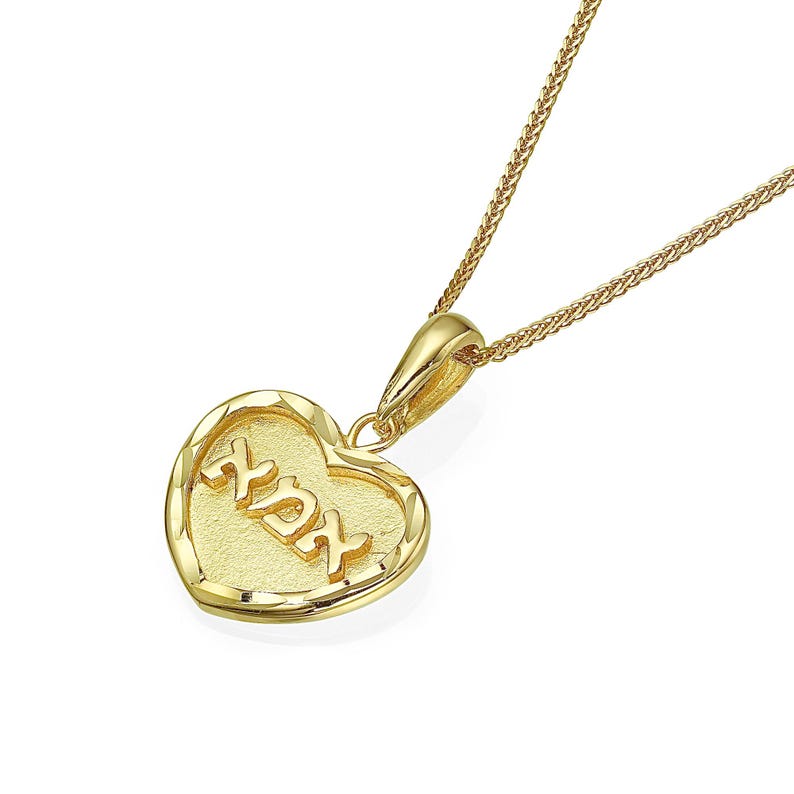 Puede incluir: Un collar con colgante en forma de coraz&oacute;n de tono dorado con un borde texturizado y las letras hebreas 'אני לדודי' en relieve en el centro. El colgante est&aacute; unido a una delicada cadena de trigo de tono dorado con acabado pulido y reasa.