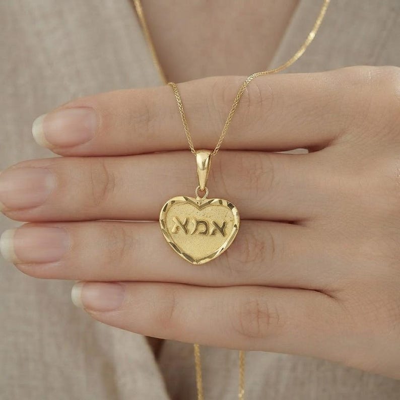 Puede incluir: Un colgante de oro en forma de coraz&oacute;n con una superficie texturizada y la palabra hebrea "אמא" grabada en el centro. El colgante cuelga de una delicada cadena de oro, mostrando un borde pulido y un dise&ntilde;o cl&aacute;sico y sencillo.