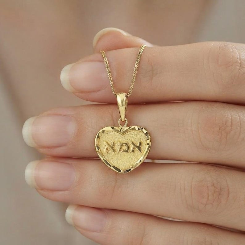 Puede incluir: Un collar con colgante en forma de coraz&oacute;n de oro con un borde texturizado y la palabra hebrea para madre, 'אמא', grabada en el centro. El colgante cuelga de una delicada cadena de oro, mostrando un acabado pulido en las letras y una superficie mate.