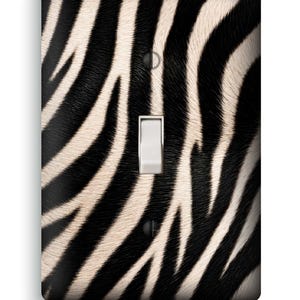 May include: A rectangular light switch wall plate featuring a black and white zebra stripe pattern. The plate has a centered white toggle switch and two black screws positioned at the top and bottom for mounting to a wall electrical box.