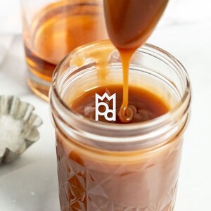 May include: A glass jar filled with a thick, golden caramel sauce. Caramel is being poured from a spoon into the jar.