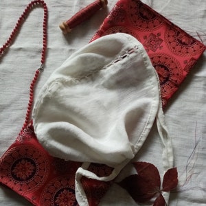 May include: White linen bonnet with a lace trim and a red and gold patterned fabric underneath. The bonnet has a long tie that can be tied under the chin.