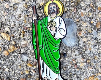 May include: A green and white enamel pin depicting Saint Patrick, a religious figure, holding a staff. He is wearing a green robe and has a halo above his head.