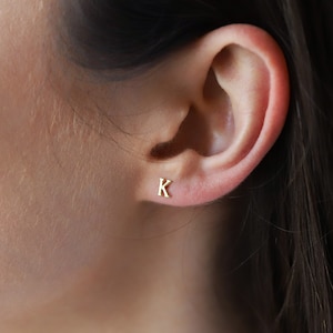 May include: A small, gold-colored stud earring shaped like the capital letter K. The earring is positioned on the lower part of the earlobe, featuring a polished metallic finish and a simple, clean-lined serif font design for the letter.
