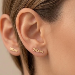 May include: Gold-colored stud earrings, one spelling "Mary" in cursive script. The other earring spells "Mary" as well. The earrings are worn on a person's ear.