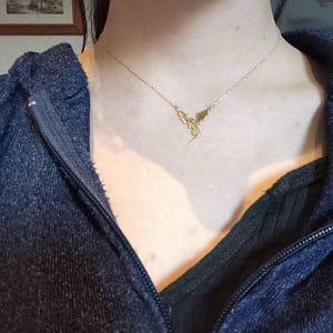 A gold-toned necklace featuring a delicate chain and a small, winged dragon pendant. The pendant is centered on the collarbone, showcasing detailed wings and a slender tail, suspended from a thin, polished metal link chain.