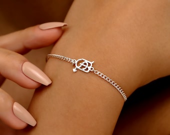 Op de afbeelding: Een zilveren armband met een schakelketting ontwerp. De armband heeft een decoratief middelpunt met een pijl en de letters "G" en "B". De armband zit om de pols van een persoon.
