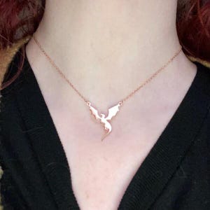 A rose gold-toned necklace featuring a delicate, flat metal pendant shaped like a flying dragon with outstretched wings. The pendant is suspended from a thin, matching rose gold-toned chain that rests against the collarbone area.