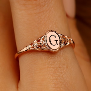A rose gold ring with a delicate filigree design. The ring features a round center stone with the letter 'G' engraved on it.