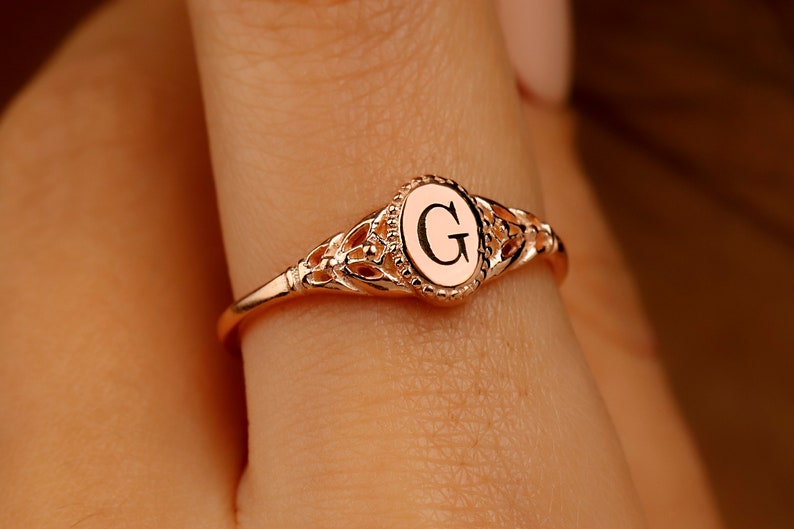 A rose gold ring with a delicate filigree design. The ring features a round center stone with the letter 'G' engraved on it.