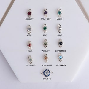 A white display board with silver charms with different colored gemstones. The charms are labeled with the months of the year, January through December, and 'Evil Eye'.