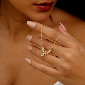 A gold-toned necklace featuring a delicate chain and a central pendant shaped like a flying dragon with outstretched wings. The dragon pendant is crafted with smooth, polished surfaces and is suspended from a thin, matching gold-toned link chain.