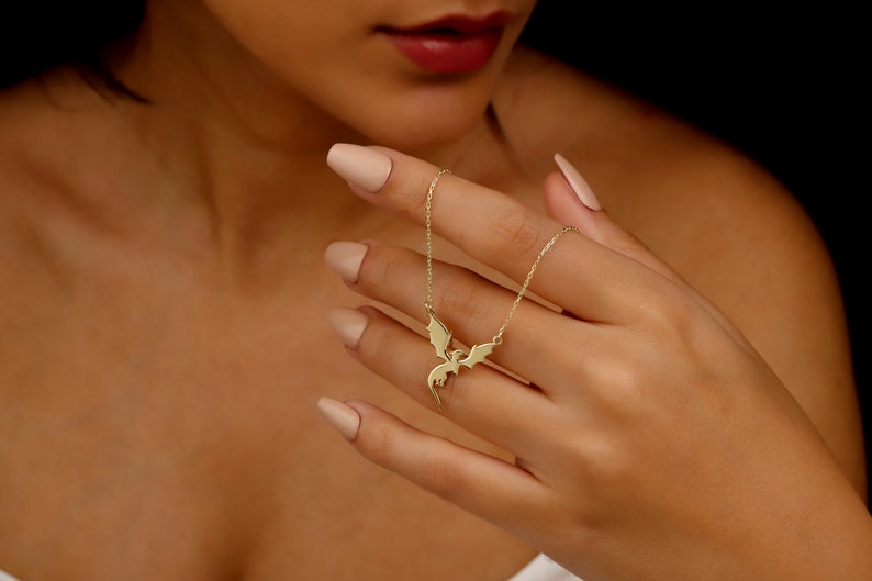 A gold-toned necklace featuring a delicate chain and a central pendant shaped like a flying dragon with outstretched wings. The dragon pendant is crafted with smooth, polished surfaces and is suspended from a thin, matching gold-toned link chain.