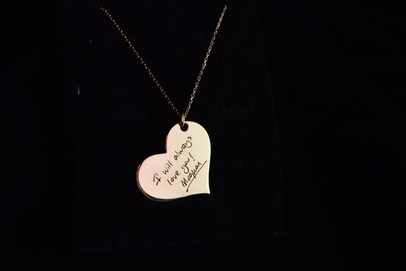 A gold heart-shaped pendant necklace with a handwritten message that reads 'I will always love you! Meghan'.