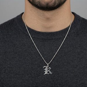 Puede incluir: Un collar con colgante plateado con la letra "K" en fuente gótica. El colgante cuelga de una fina cadena. La persona lleva un jersey gris oscuro.