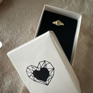 A gold ring with an oval-shaped setting featuring the letter 'E' in black lettering in a NecklaceDreamWorld`s gift box.