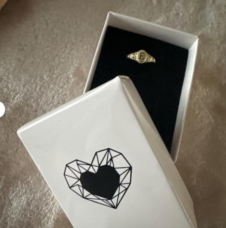 A gold ring with an oval-shaped setting featuring the letter 'E' in black lettering in a NecklaceDreamWorld`s gift box.