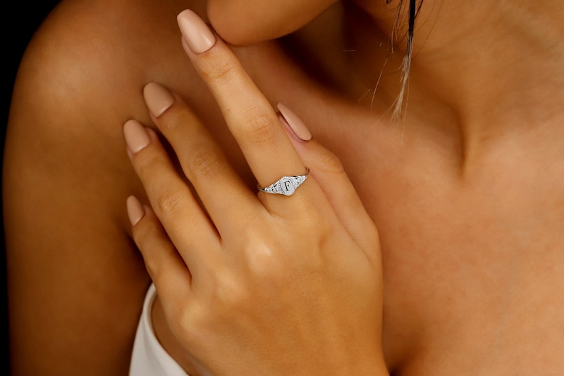 A silver signet ring with a raised letter 'F' in the center. The ring is set with small, clear stones around the letter.
