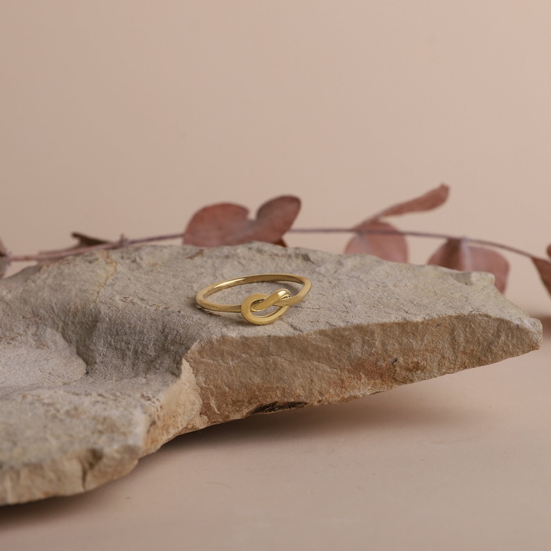 A gold-colored ring with a knot design. The ring is sitting on a light brown rock.