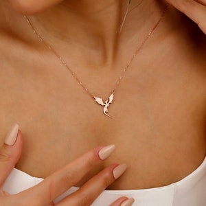 A rose gold-toned necklace featuring a delicate, flat pendant shaped like a flying dragon with outstretched wings. The pendant is suspended from a thin, matching rose gold-toned chain that rests against the collarbone area.