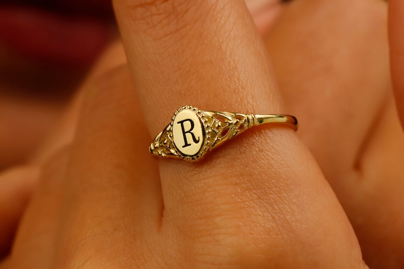 A gold ring with an oval-shaped setting featuring the letter 'R' in black lettering.