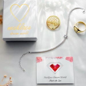 A white gift box with a gold heart and the text "necklace dream world," a silver-tone tennis necklace, a gold-tone tree of life pendant, a gold-tone signet ring, and a white card featuring a red heart diamond graphic and text.
