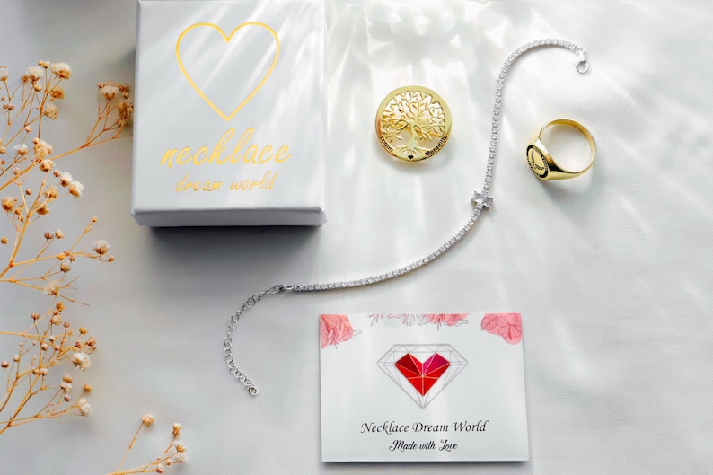 A white gift box with a gold heart and the text "necklace dream world," a silver-tone tennis necklace, a gold-tone tree of life pendant, a gold-tone signet ring, and a white card featuring a red heart diamond graphic and text.
