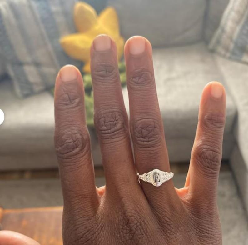 A silver signet ring with a raised letter 'F' in the center. The ring is set with small, clear stones around the letter.