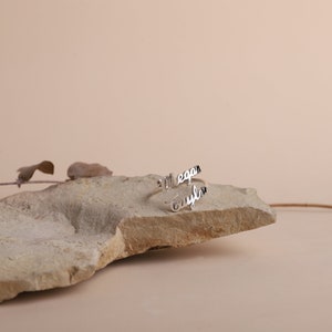 A silver ring with the names 'Megan' and 'Taylor' engraved on it. The ring is sitting on a light brown rock.