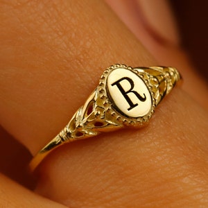 A gold ring with an oval-shaped setting featuring the letter 'R' in black lettering.