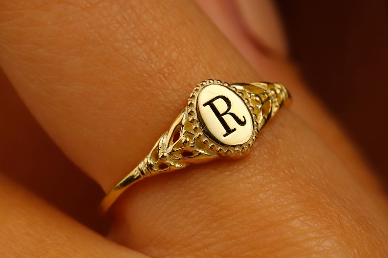 A gold ring with an oval-shaped setting featuring the letter 'R' in black lettering.