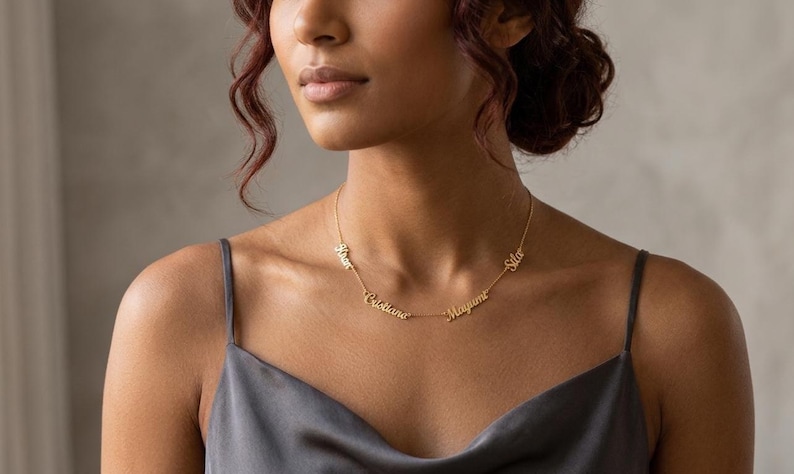 Puede incluir: Un collar dorado con m&uacute;ltiples colgantes con nombres. El collar lo lleva una persona con un top de sat&eacute;n gris. Los nombres est&aacute;n escritos en una fuente cursiva.