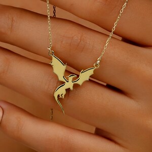 A gold-toned necklace featuring a delicate pendant shaped like a flying dragon with outstretched wings. The pendant is suspended from a thin, matching gold-toned chain, creating a simple and elegant piece of jewelry for the neck.