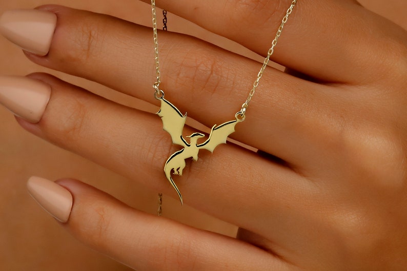 A gold-toned necklace featuring a delicate pendant shaped like a flying dragon with outstretched wings. The pendant is suspended from a thin, matching gold-toned chain, creating a simple and elegant piece of jewelry for the neck.