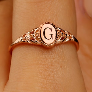 A rose gold ring with a delicate filigree design. The ring has a center oval with the letter 'G' engraved inside.