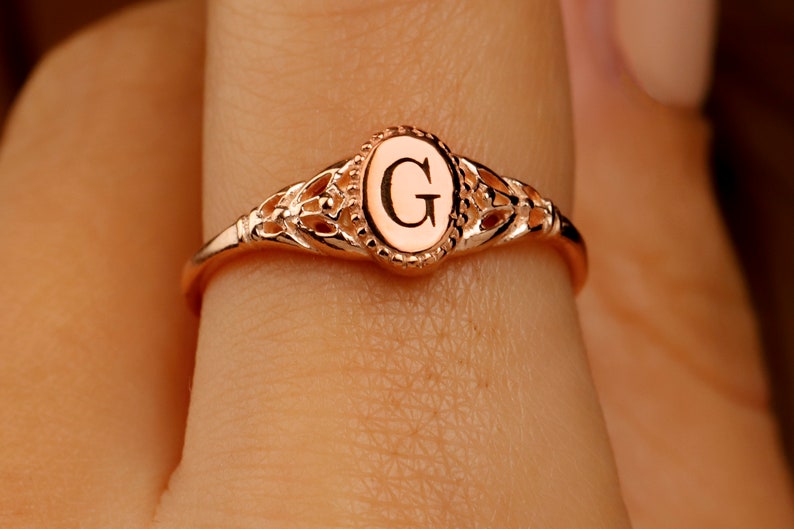 A rose gold ring with a delicate filigree design. The ring has a center oval with the letter 'G' engraved inside.