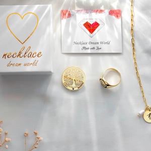 May include: A white gift box with a gold heart and the text necklace dream world, a card with a red heart logo and text, a gold tree of life circular pendant, a gold signet ring, and a gold chain necklace with a letter B pendant and a small pearl.