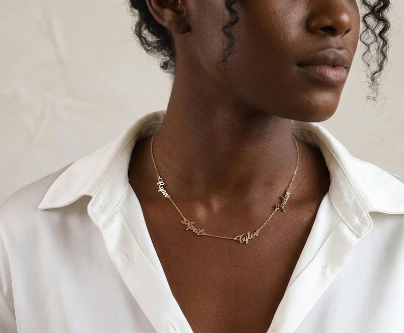 Puede incluir: Un collar dorado con los nombres "Ryan", "April", "Tyler" y "Jordan" en escritura cursiva. El collar se muestra sobre una camisa blanca, resaltando la joyer&iacute;a personalizada.