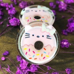Pode incluir: Dois brincos em forma de donut com rosto de gato rosa e branco e um laço branco no topo. Os brincos são feitos de resina transparente e têm um acabamento brilhante.